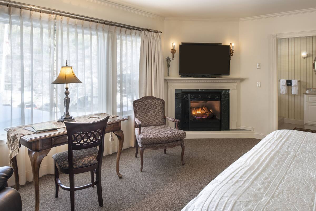 A hotel room with a fireplace, a desk with a lamp, a comfortable chair, and a neatly made bed.