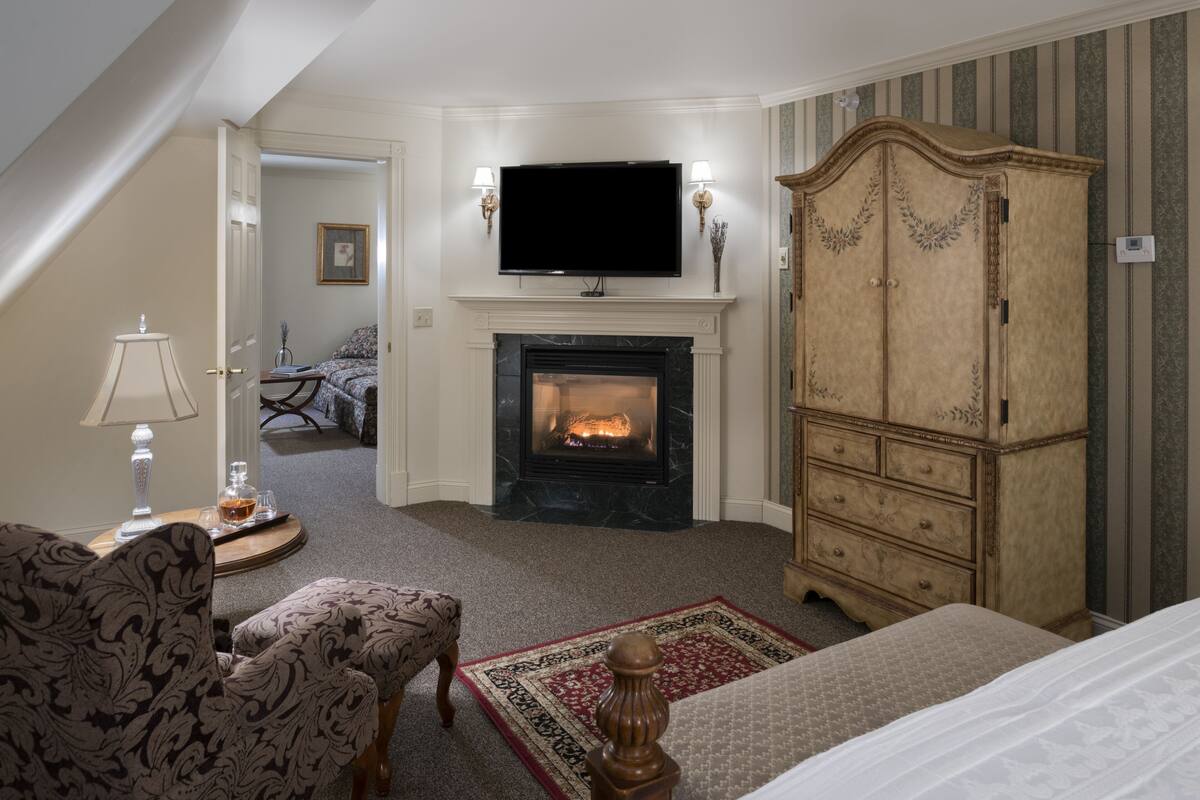 A hotel room with a fireplace, a television mounted above it, a large wooden wardrobe, a bed, a patterned armchair, and a small table with a lamp.