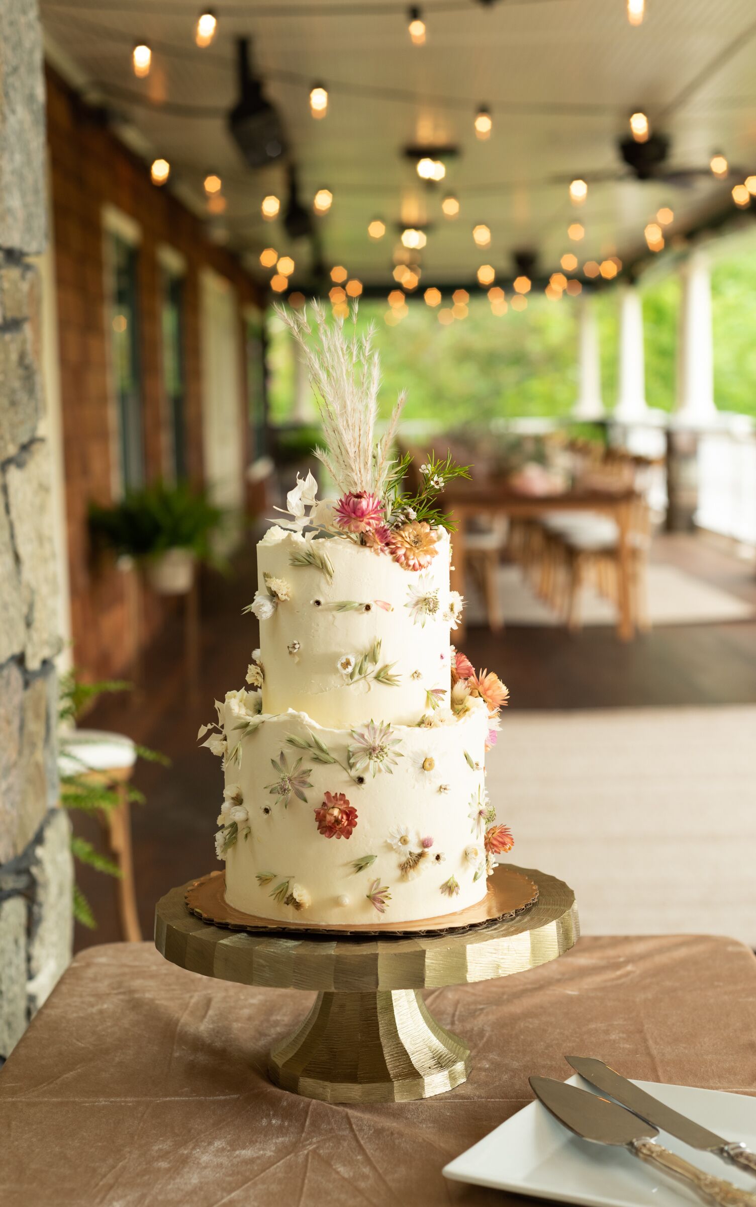 A multi-tiered cake with floral decorations on a wooden stand, placed on a table in a covered outdoor area with string lights in the background.