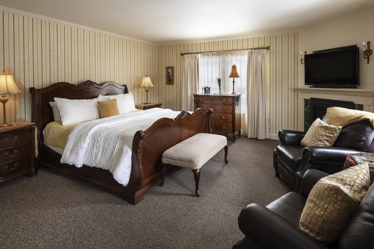 A bedroom with a large bed, a wooden dresser, a leather armchair, a television, and a fireplace.