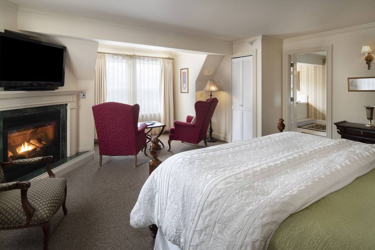 A hotel room with a fireplace, a bed, two armchairs, a television, and a mirror.