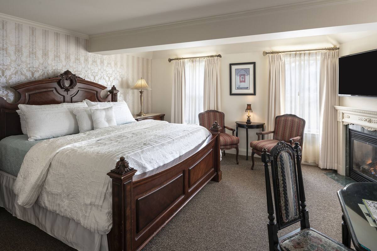 A hotel room with a large bed, two armchairs, a small table, a fireplace, and a television.