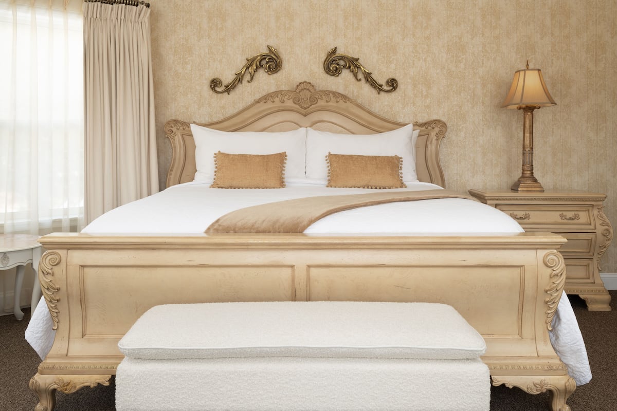 A large bed with a headboard, two pillows, a bedside table with a lamp, and a window with curtains.