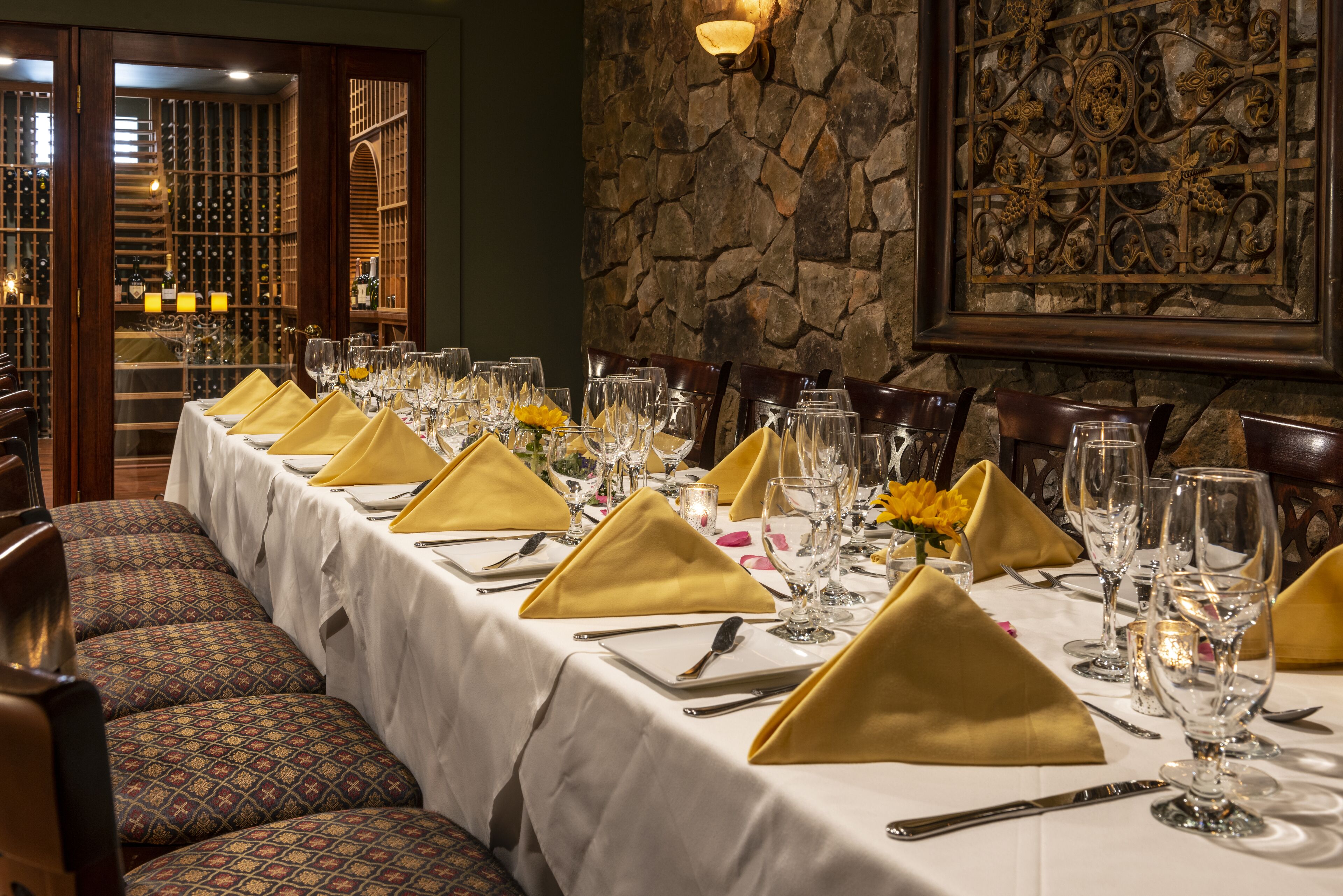 A long dining table set with gold napkins, wine glasses, and cutlery, surrounded by patterned chairs.