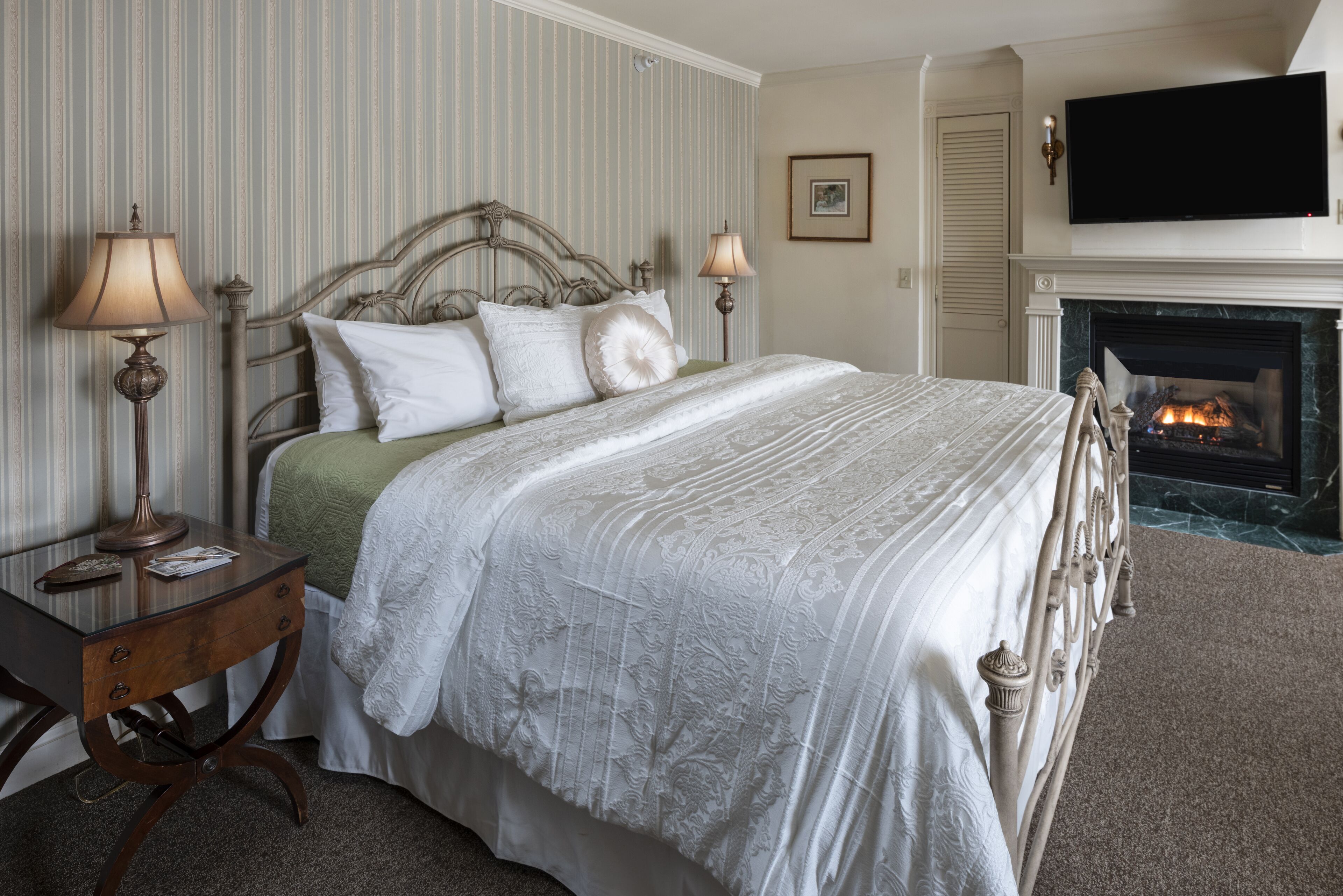 A neatly arranged bedroom with a large bed, two bedside lamps, a fireplace, and a television mounted above it.