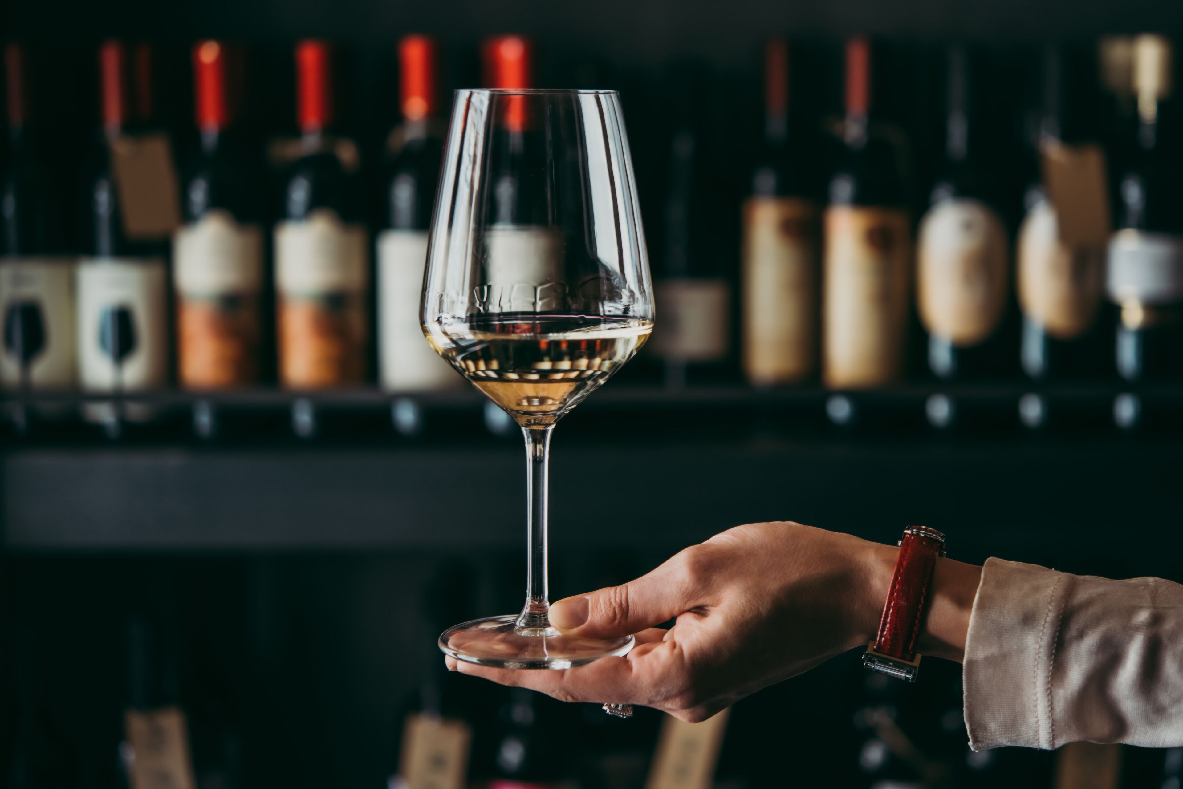 Une main tenant un verre à vin, avec un arrière-plan flou composé de bouteilles de vin.