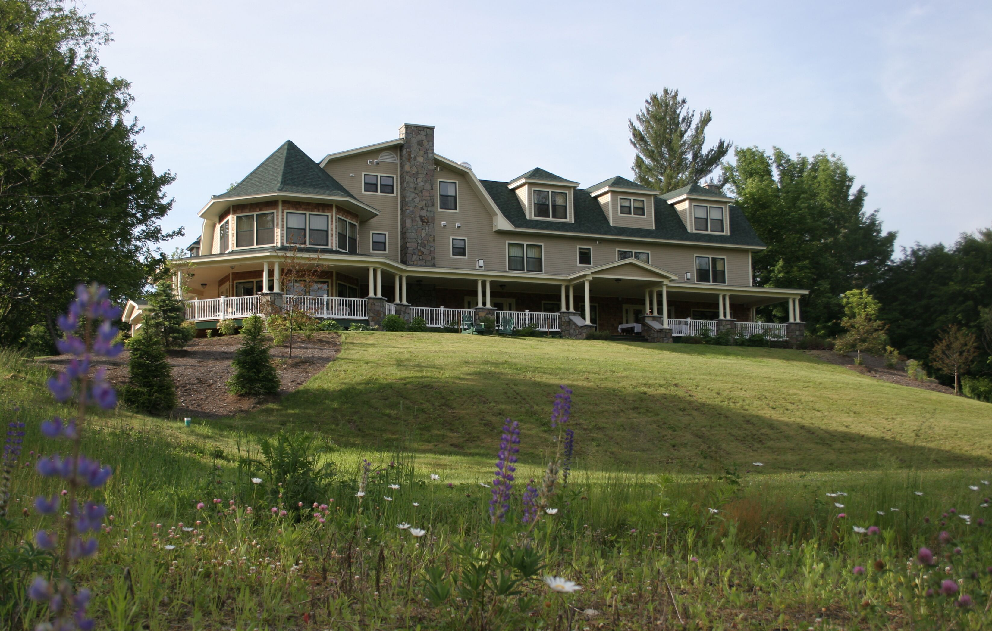 A large, multi-story house with a stone chimney, multiple balconies, and a well-maintained lawn with wildflowers.