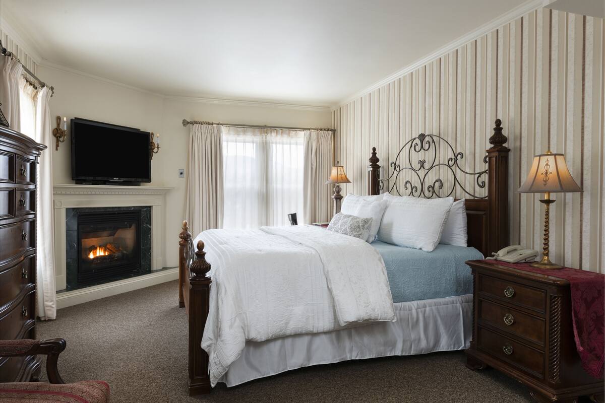 A bedroom with a fireplace, a bed with white bedding, two bedside lamps, and a wooden dresser.
