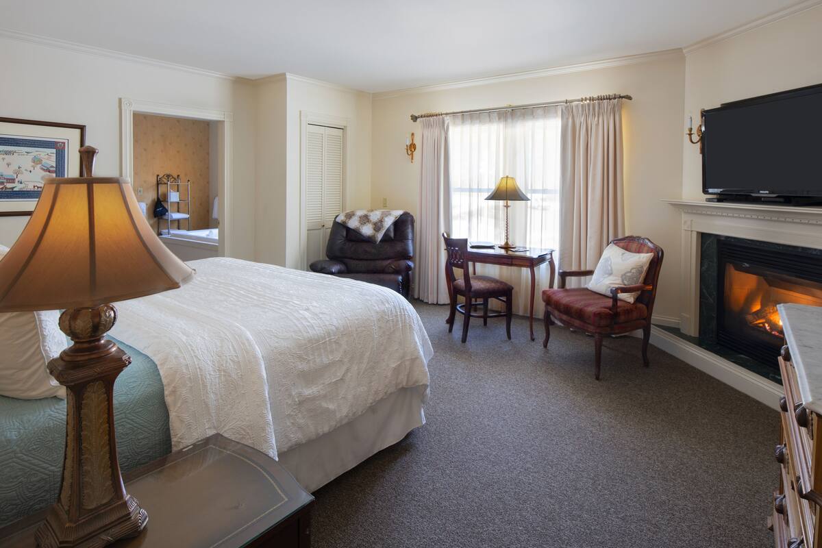 A hotel room with a bed, a desk, a chair, a television, and a fireplace.