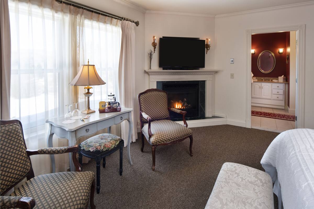 A hotel room with a fireplace, a television, a desk with a lamp, and a seating area with chairs and a sofa.