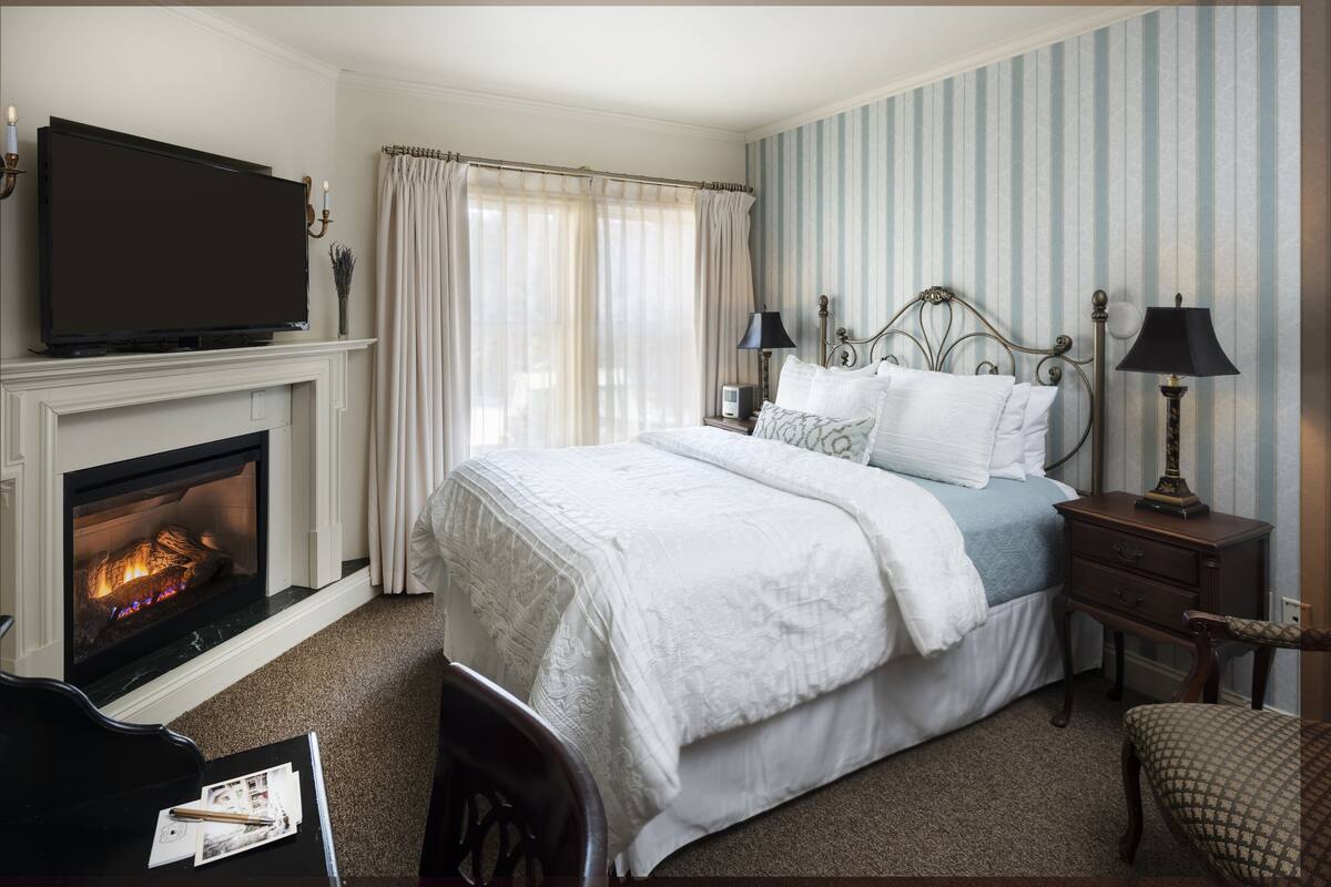 A hotel room with a fireplace, a bed with white bedding, a television, and a nightstand with lamps.