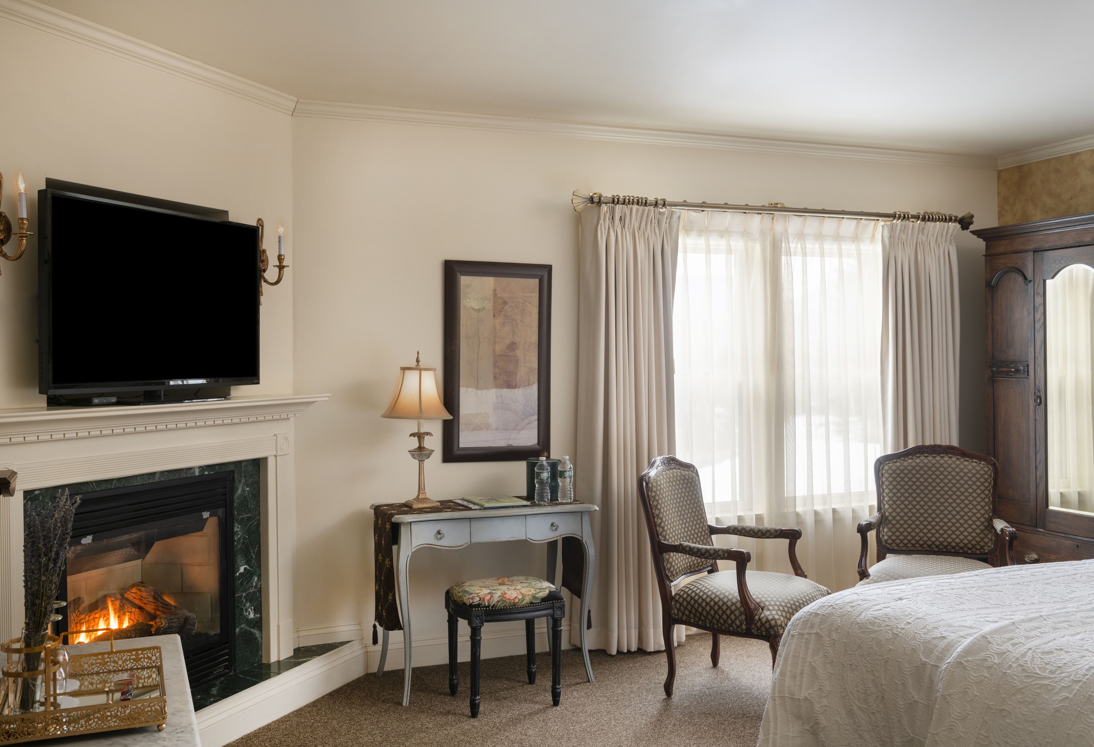 A hotel room with a fireplace, a television, a desk with a lamp, two chairs, and a bed.