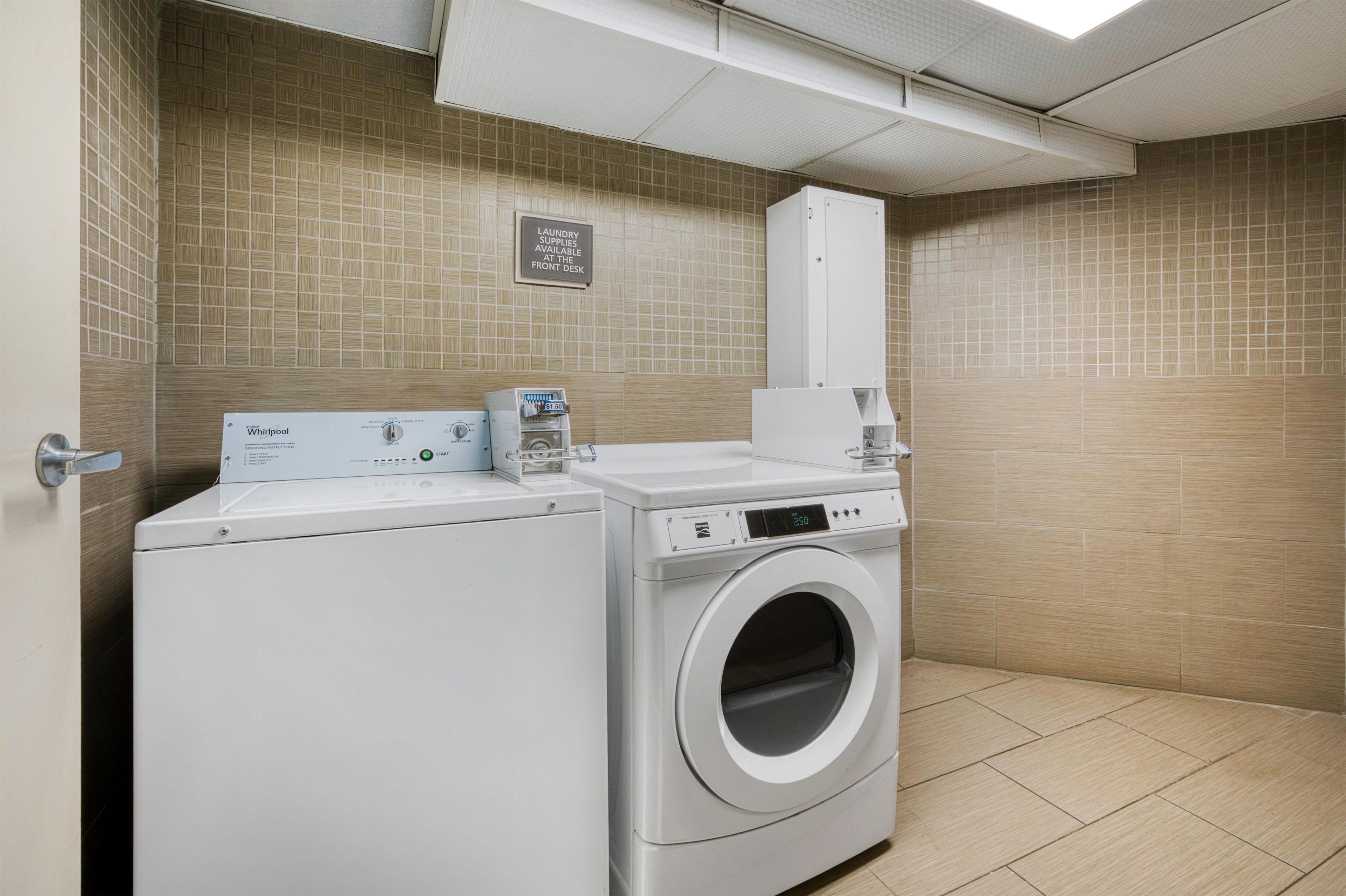 A laundry room with a Whirlpool washer and dryer, a sign indicating laundry supplies at the front desk, and a tiled wall.