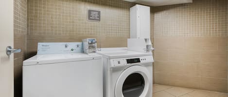 A laundry room with a Whirlpool washer and dryer, a sign indicating laundry supplies at the front desk, and a tiled wall.