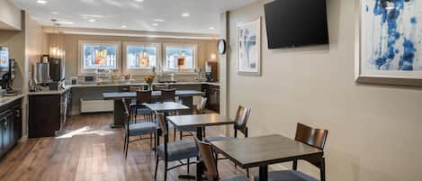 A modern dining area with tables and chairs, a kitchen in the background, and artwork on the walls.