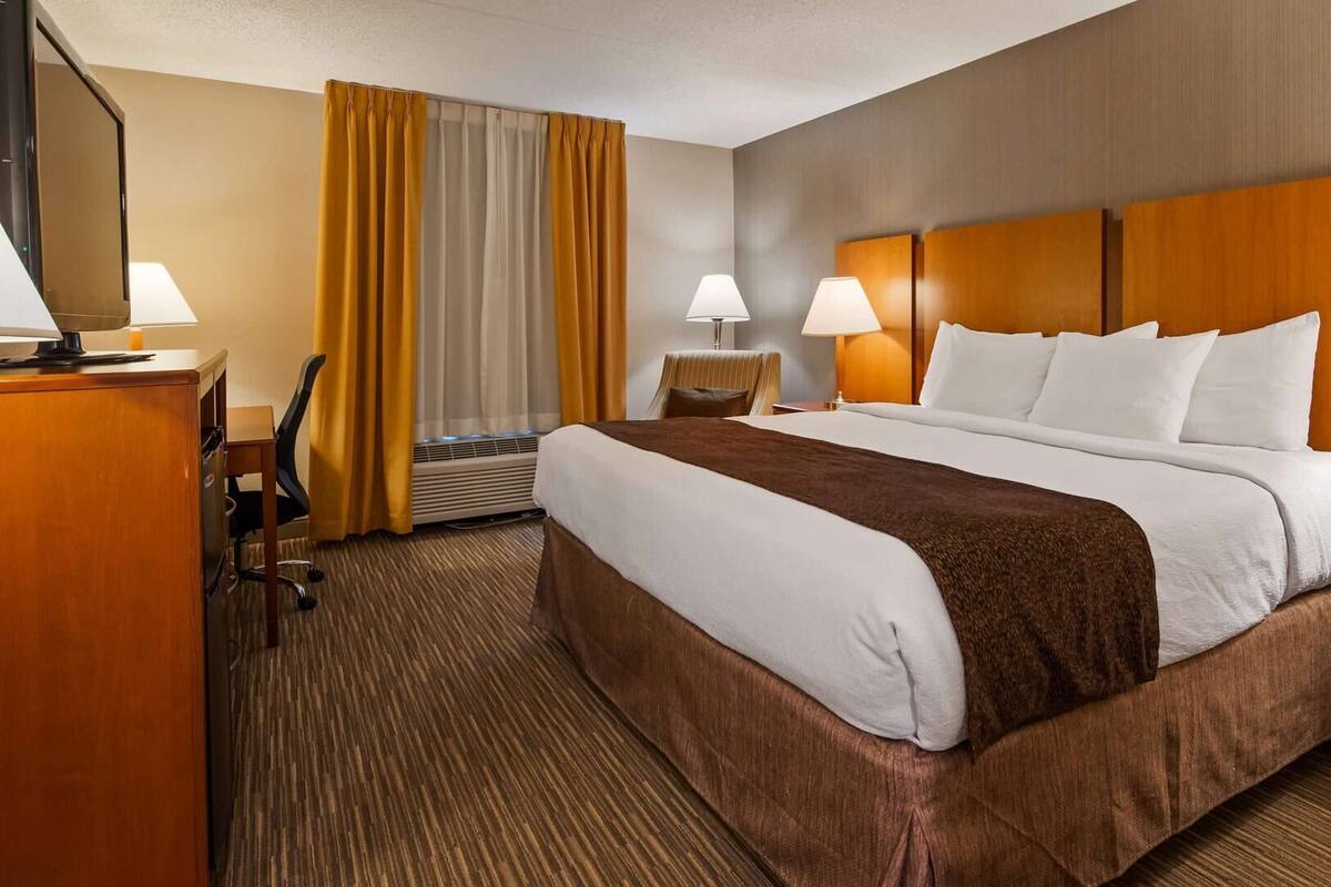 Ein Hotelzimmer mit einem großen Bett, zwei Nachttischen mit Lampen, einem Schreibtisch mit Stuhl und einem Fenster mit Vorhängen.