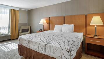 Ein Hotelzimmer mit einem großen Bett, zwei Nachttischlampen, einem Sessel, einem Schreibtisch und einem Fenster mit Vorhängen.