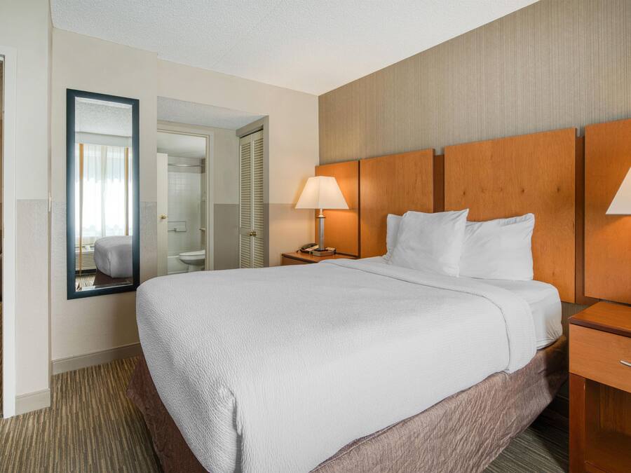 A hotel room with a large bed, a bedside table with a lamp, a mirror, and a view of the bathroom.
