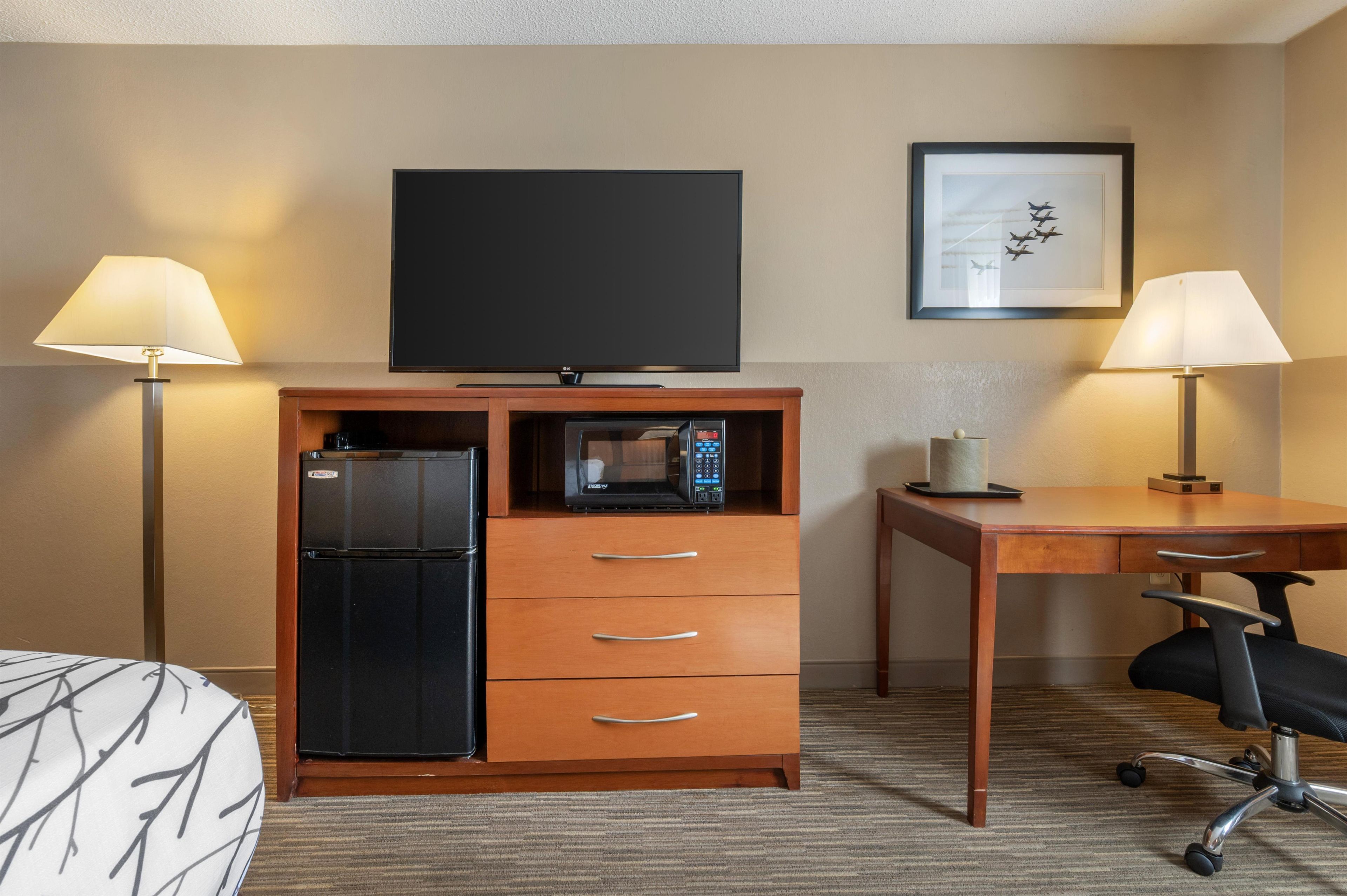 A hotel room with a flat-screen TV, a minibar, a desk with a chair, and two lamps.