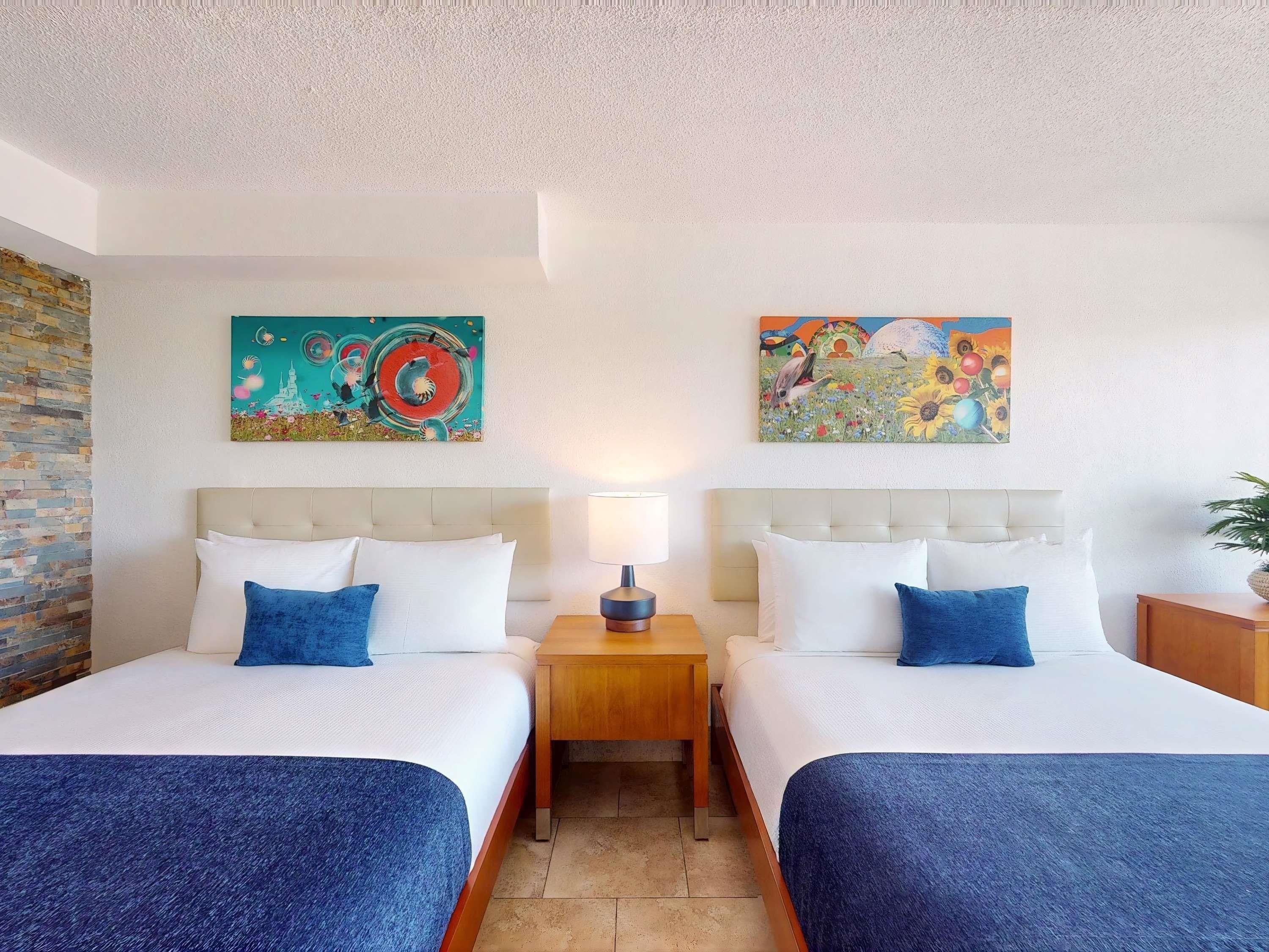 Habitación de hotel con dos camas, una mesita de noche, una lámpara y dos cuadros en la pared.