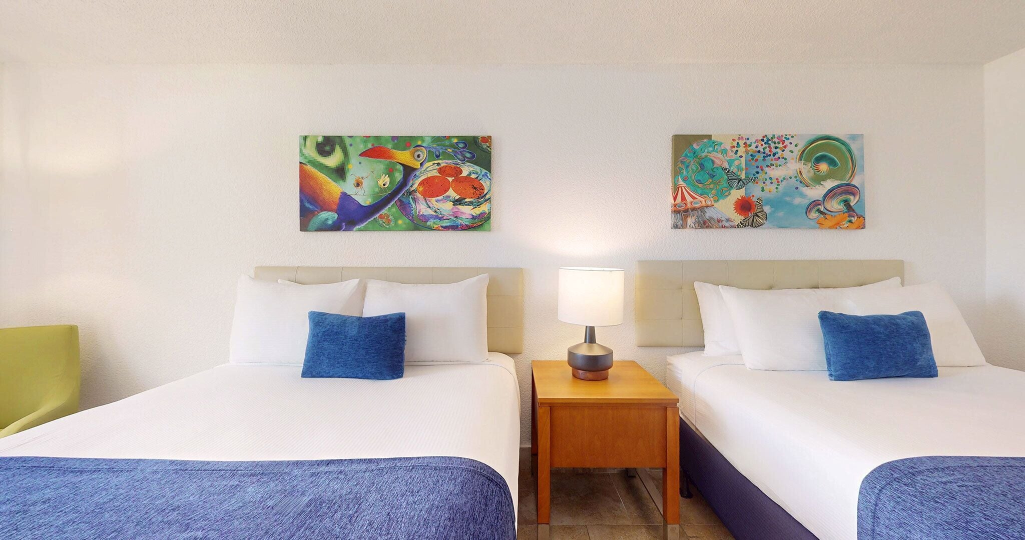 Camera d'albergo con due letti, ciascuno con biancheria da letto bianca e cuscini blu. Un comodino in legno posizionato tra i letti ospita una lampada. Le pareti sono decorate con dipinti astratti colorati.