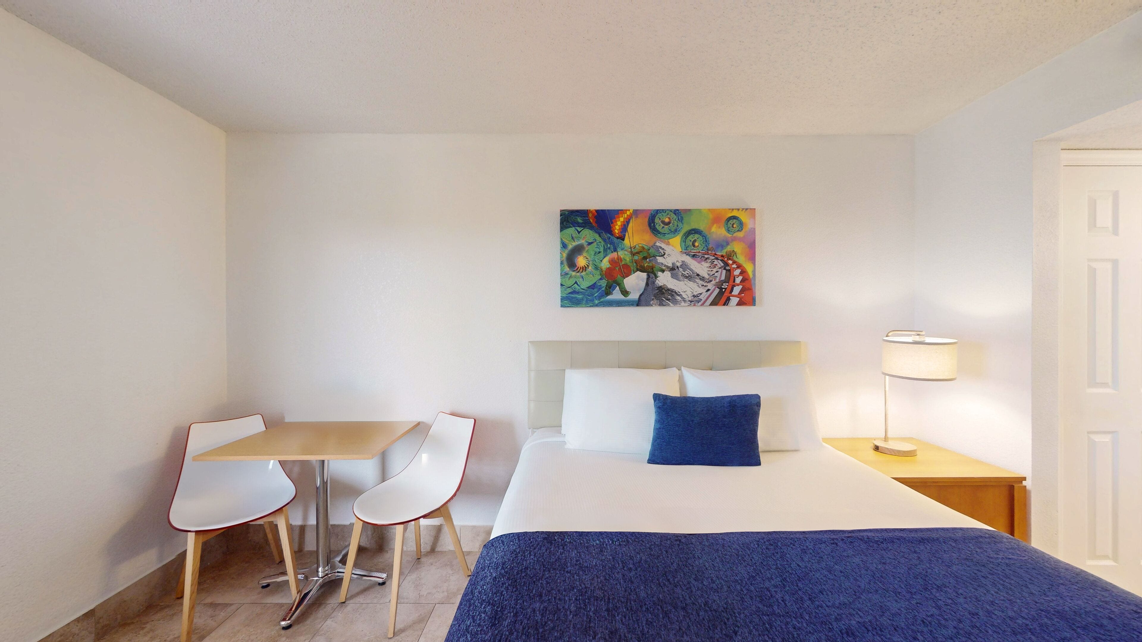 Une chambre d’hôtel avec un lit, un bureau, deux chaises, un tableau au mur et une table de chevet avec une lampe.