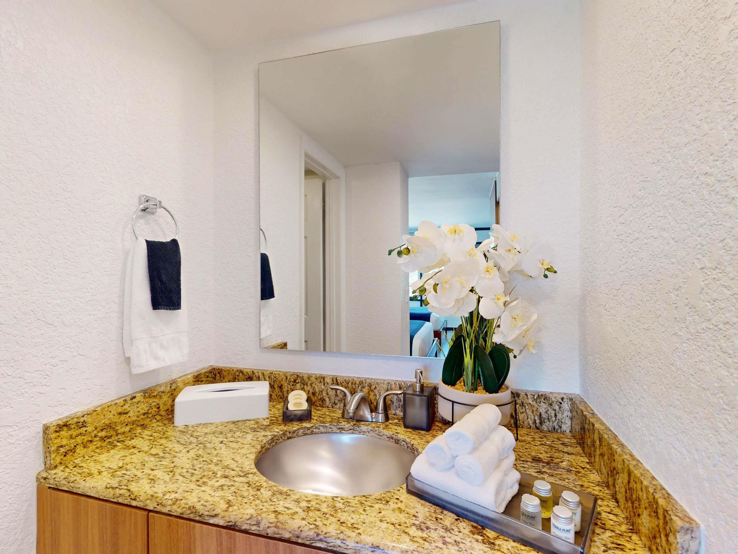 A bathroom with a granite countertop, a sink, a mirror, a towel rack, and a vase with white flowers.