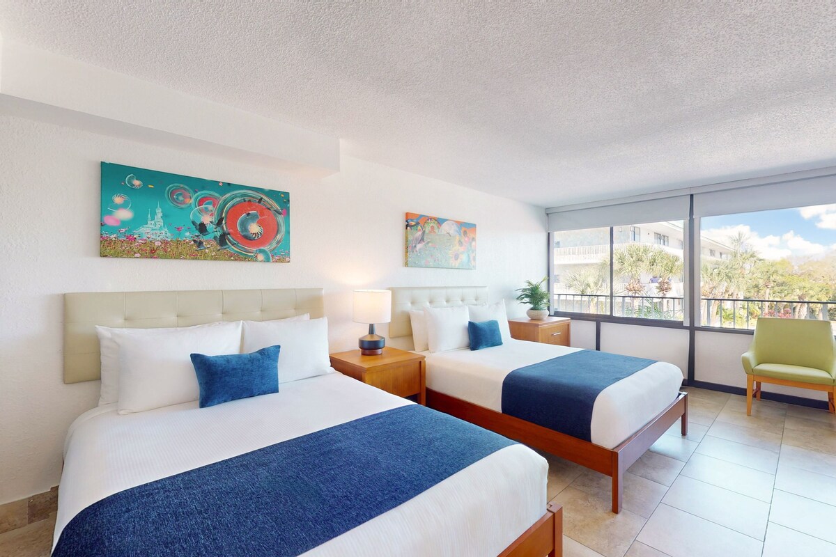 A hotel room with two beds, a large window, and artwork on the walls.