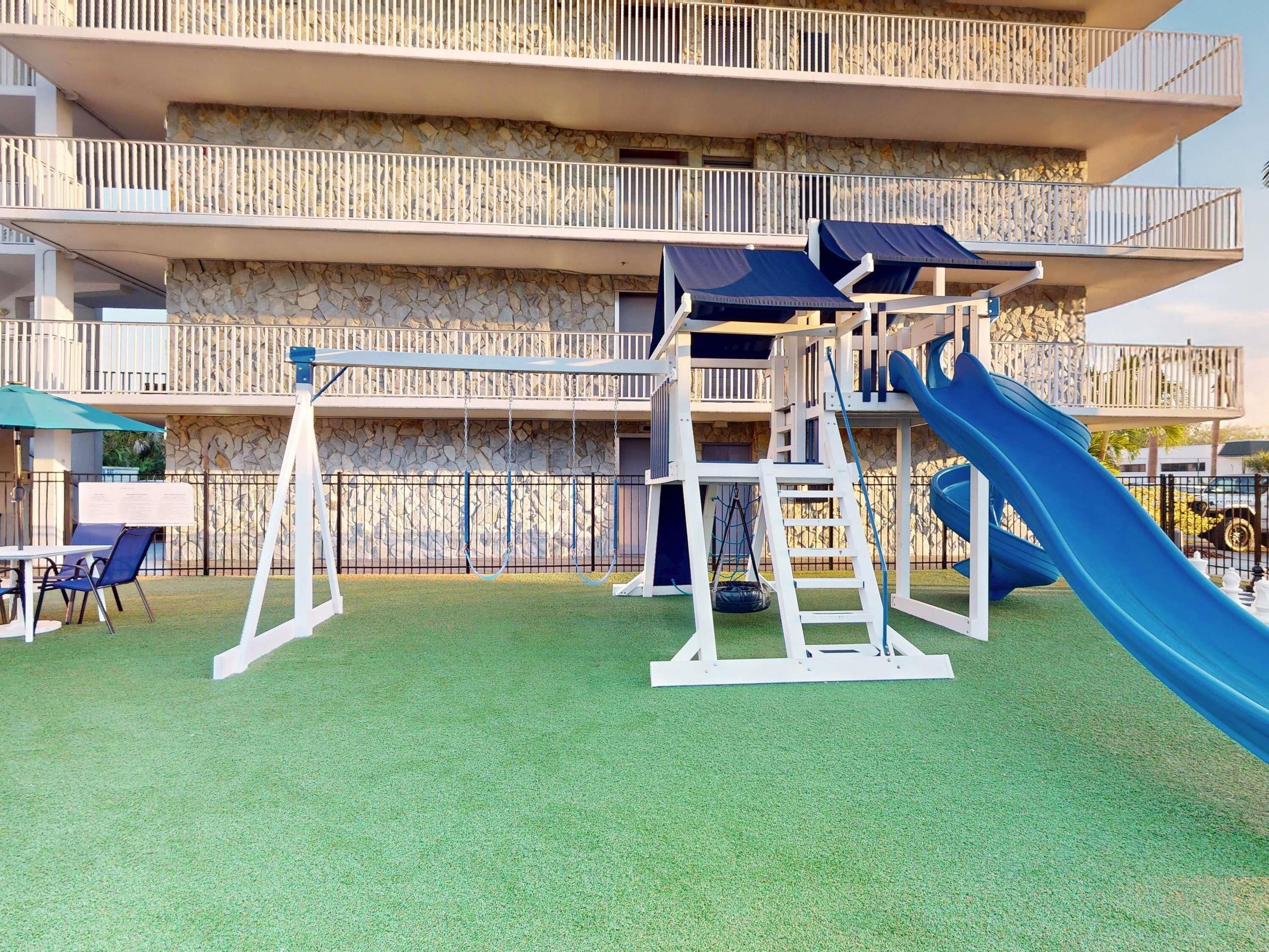 Un terrain de jeux avec des balançoires et un toboggan, entouré d’un mur en pierre et agrémenté d’un balcon.