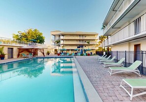 Ein Schwimmbecken mit Liegestühlen und Sonnenschirmen, ein Gebäude mit Balkonen und ein klarer Himmel.