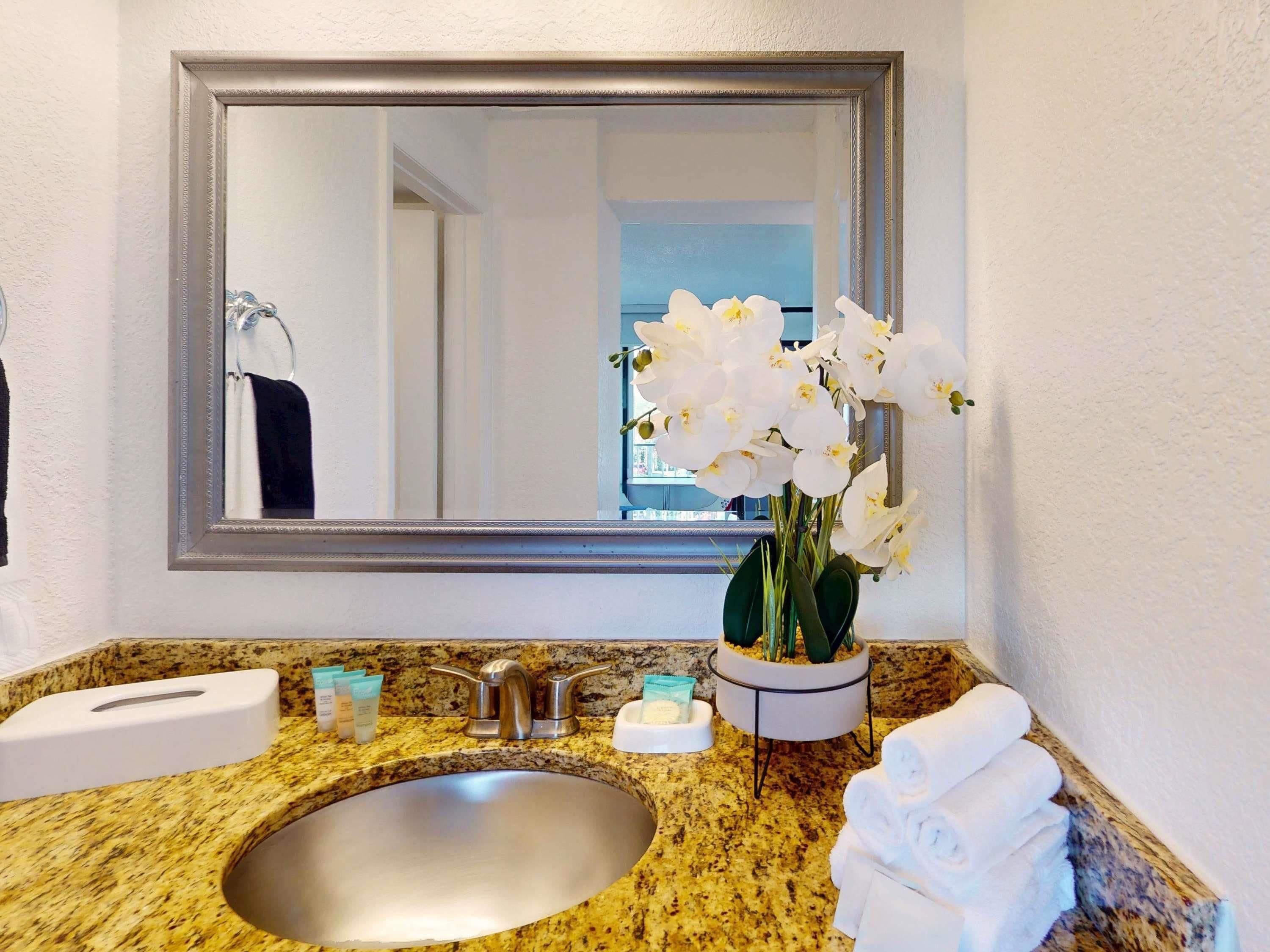 A bathroom with a granite countertop, a sink, a mirror, a towel rack, and a vase with white flowers.