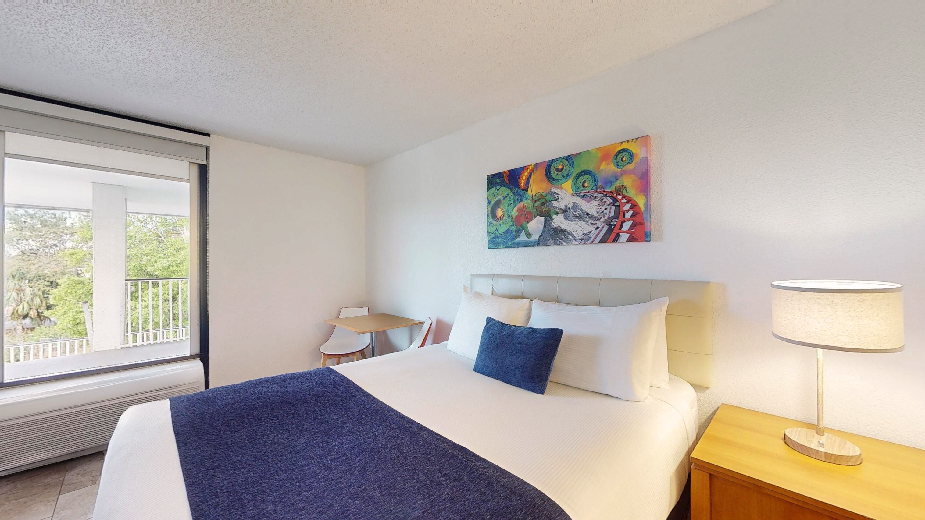 A hotel room with a bed, a desk, a chair, a window, and a painting on the wall.