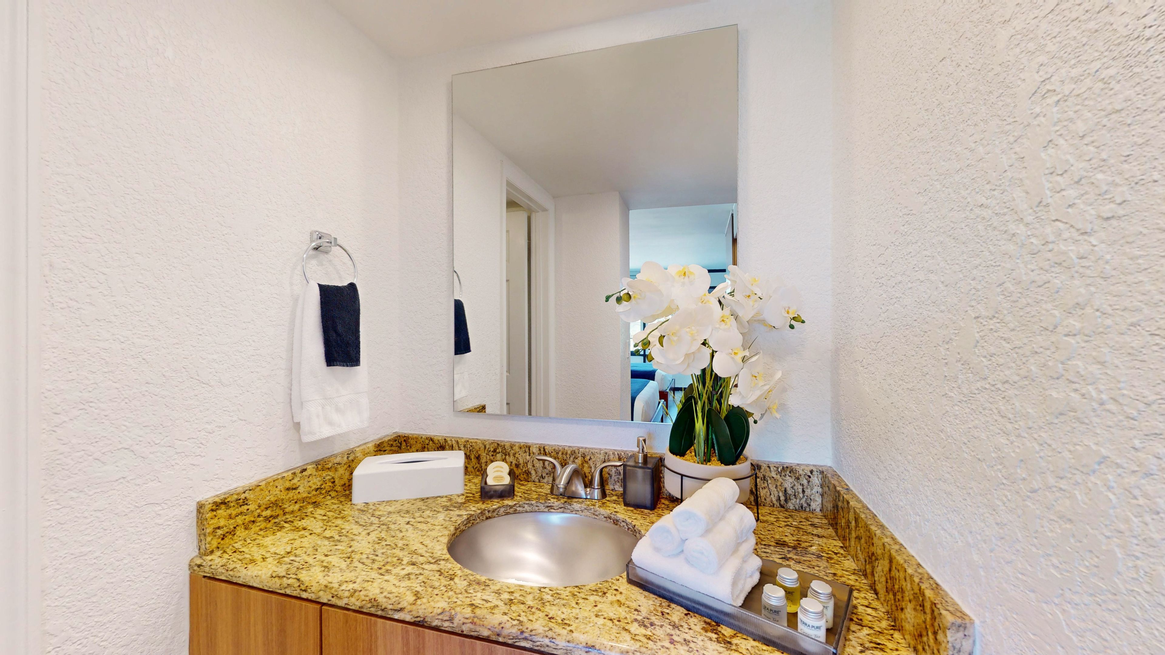A bathroom with a granite countertop, a sink, a mirror, a towel rack, and a vase with white flowers.