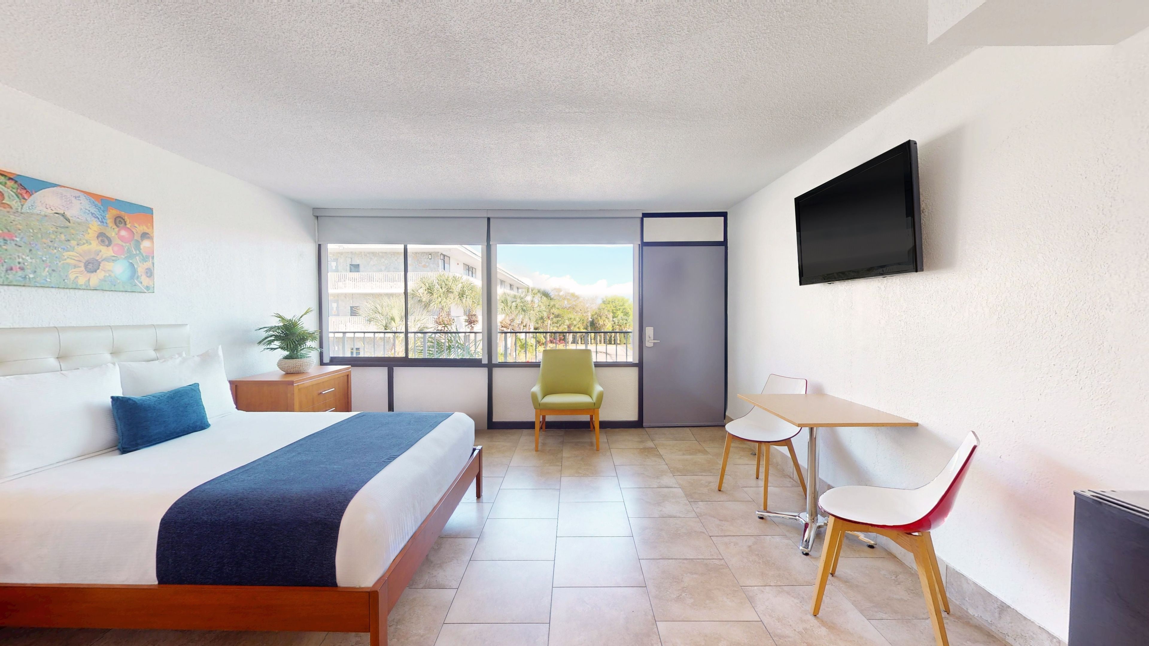 A modern hotel room with a bed, a desk, a chair, a TV, and a balcony with a view.