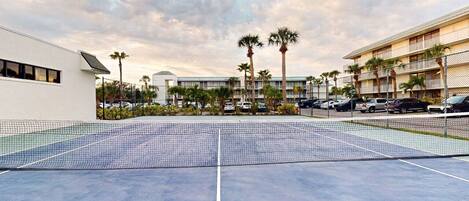 An outdoor tennis court with a net, surrounded by palm trees and a building with multiple balconies.