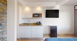 Cocina compacta con armarios blancos, microondas, cafetera y televisor montado en la pared.