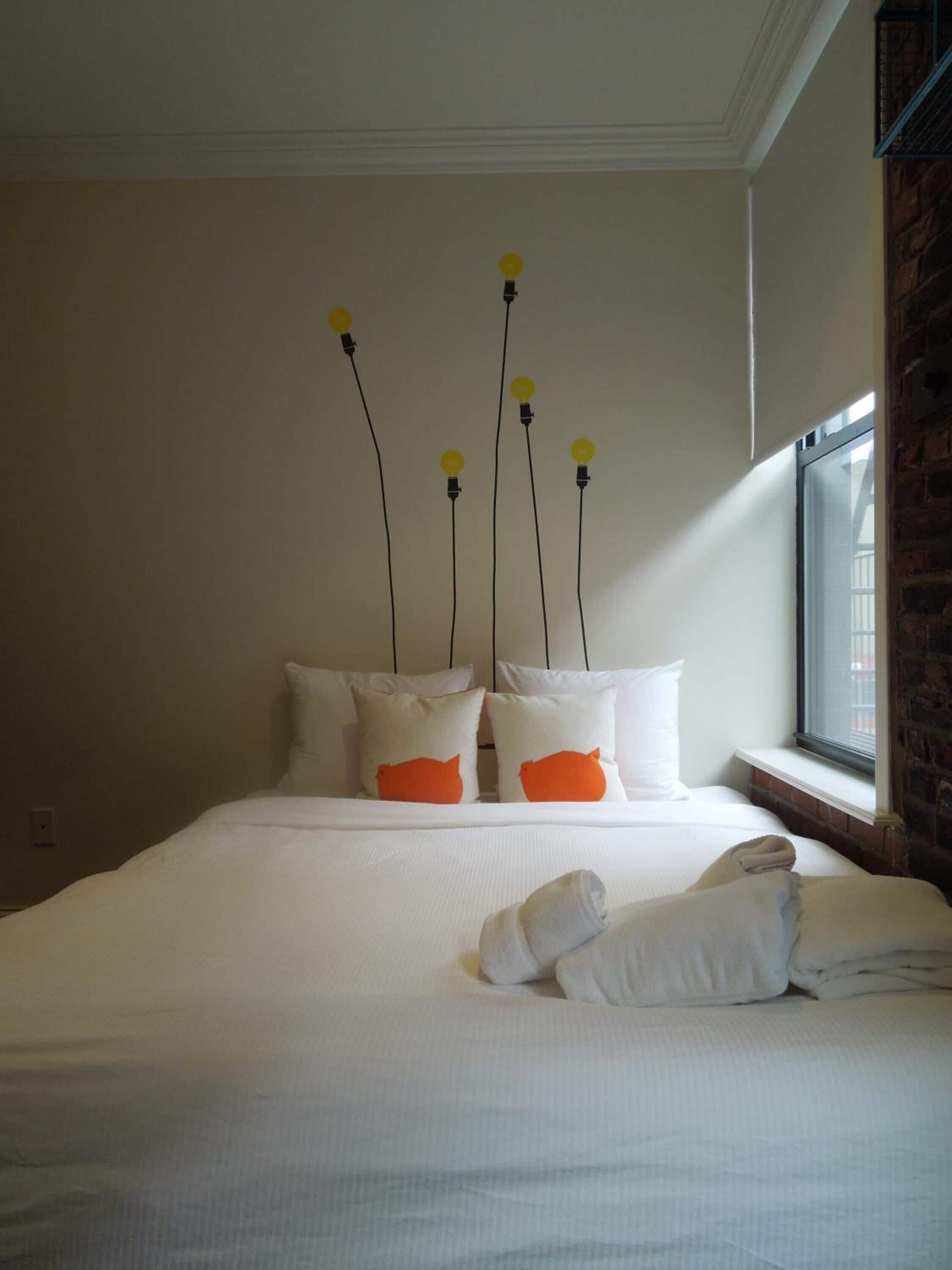 Een netjes opgemaakt bed met witte beddengoed en oranje kussens, een raam met uitzicht op een bakstenen muur, en een wandlamp met gele lampen.