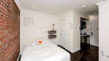 A hotel room with a brick wall, a bed with white bedding, a nightstand with a lamp, a kitchen area with a microwave, and a door leading to another room.