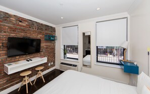 A modern hotel room with a brick wall, a flat-screen TV, a small desk with stools, a large bed, and a window with a view of the outside. - East Village Hotel (New York)