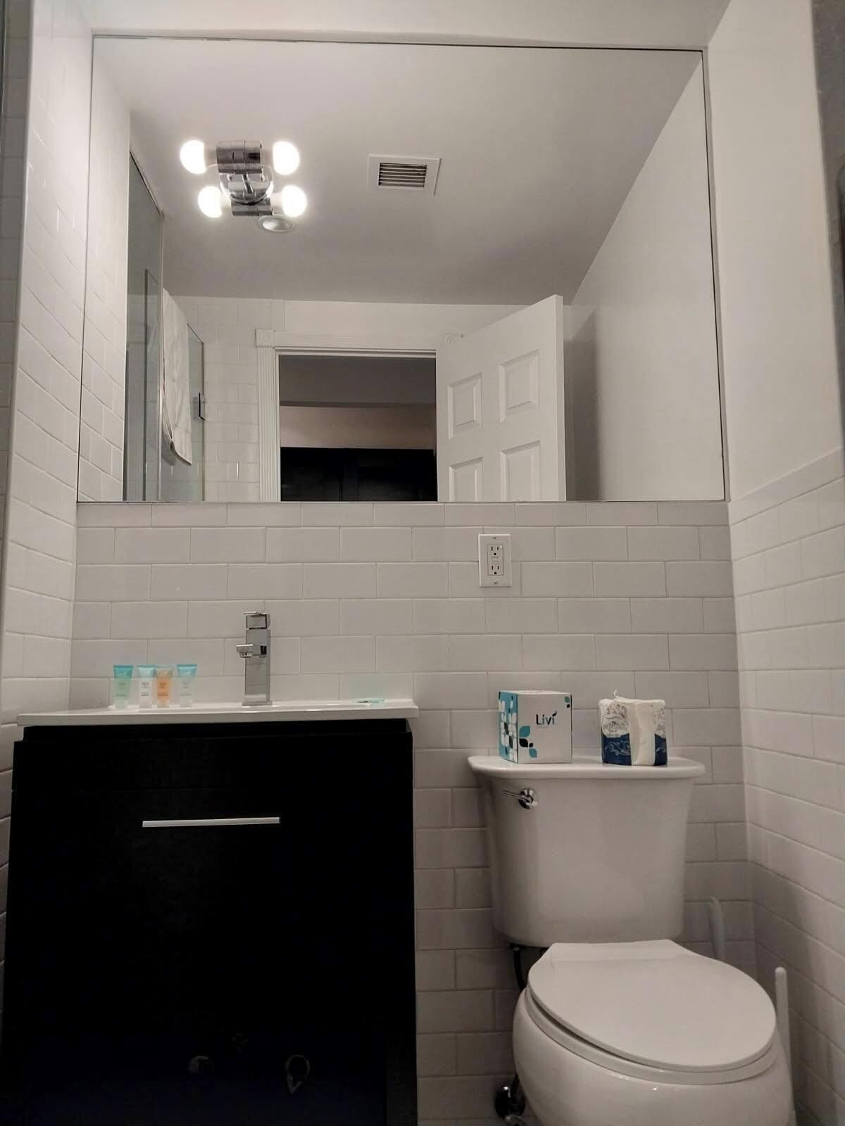 Une salle de bain avec un mur recouvert de carreaux blancs, un meuble-lavabo noir, des toilettes blanches et un miroir au-dessus du meuble-lavabo.