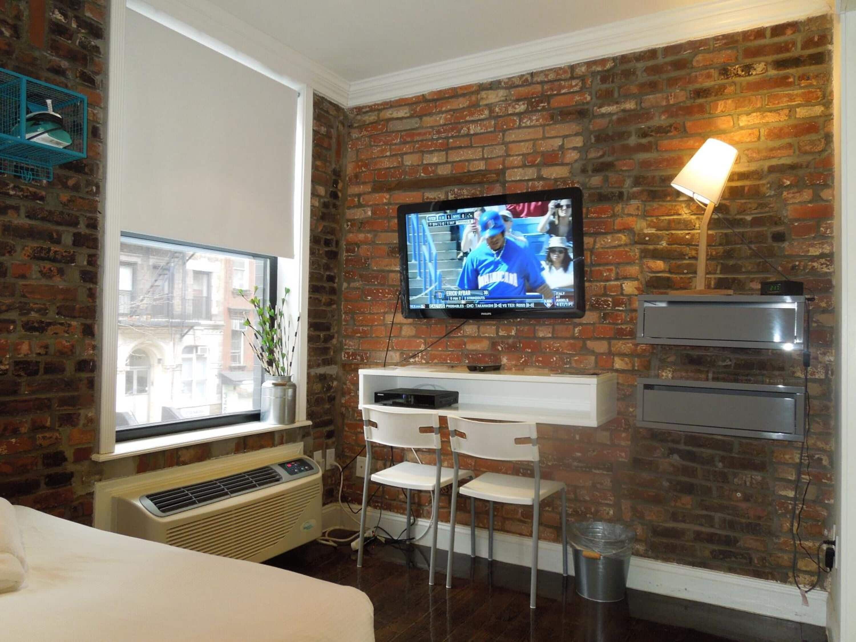 A hotel room with a brick wall, a flat-screen TV mounted above a white desk, a potted plant, and a window with a view of buildings.