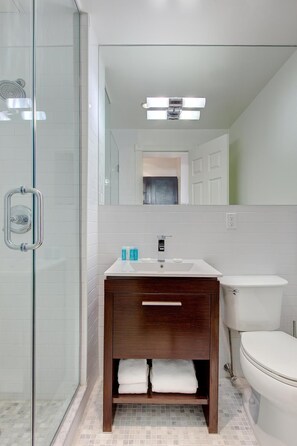 A modern bathroom with a glass shower enclosure, a white sink with a wooden vanity, and a white toilet. - East Village Hotel (New York)