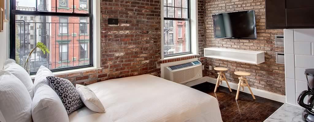 A modern hotel room with a brick wall, a large window, a bed, a TV mounted on the wall, and a small bar area.