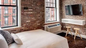 A modern hotel room with a brick wall, a large window, a bed, a TV mounted on the wall, and a small bar area.