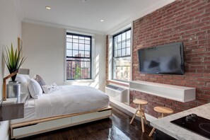 A modern bedroom with a bed, a TV mounted on a brick wall, and a window overlooking a cityscape. - East Village Hotel (New York)