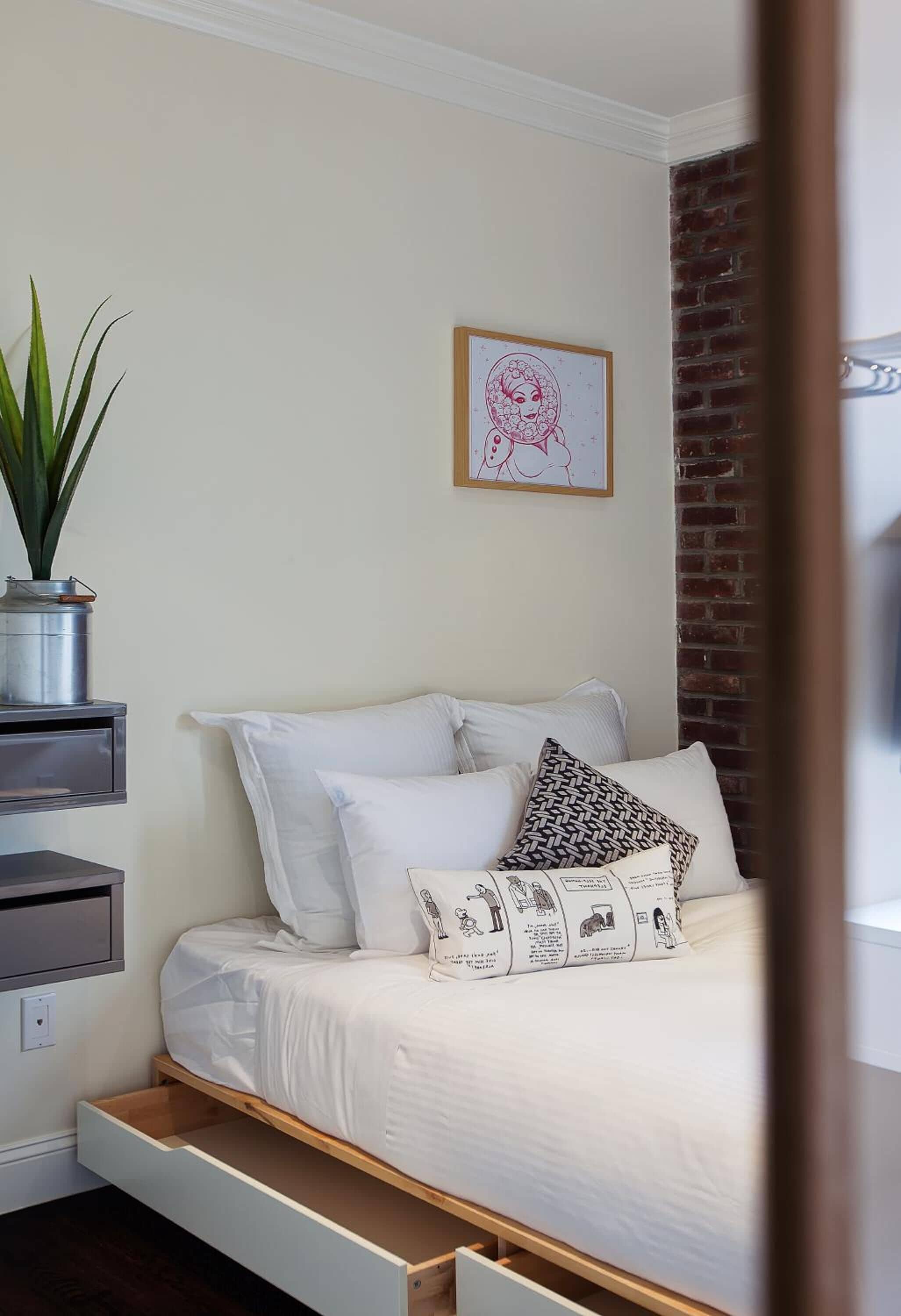 A bedroom with a bed, a nightstand, a drawer, a potted plant, and a framed picture on the wall.