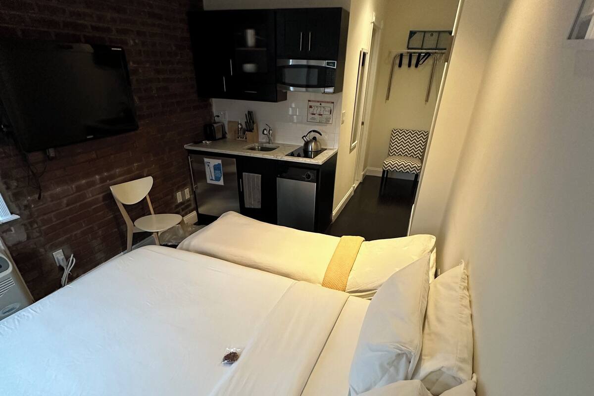 A hotel room with a large bed, a small kitchenette, a TV mounted on the wall, and a chair.