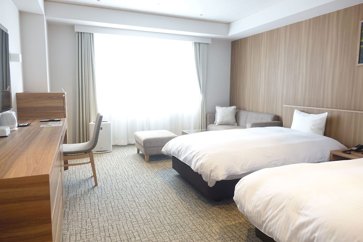 Ein Hotelzimmer mit einem Holztisch, zwei Betten, einem Sessel, einem kleinen Tisch und einem großen Fenster mit Vorhängen.