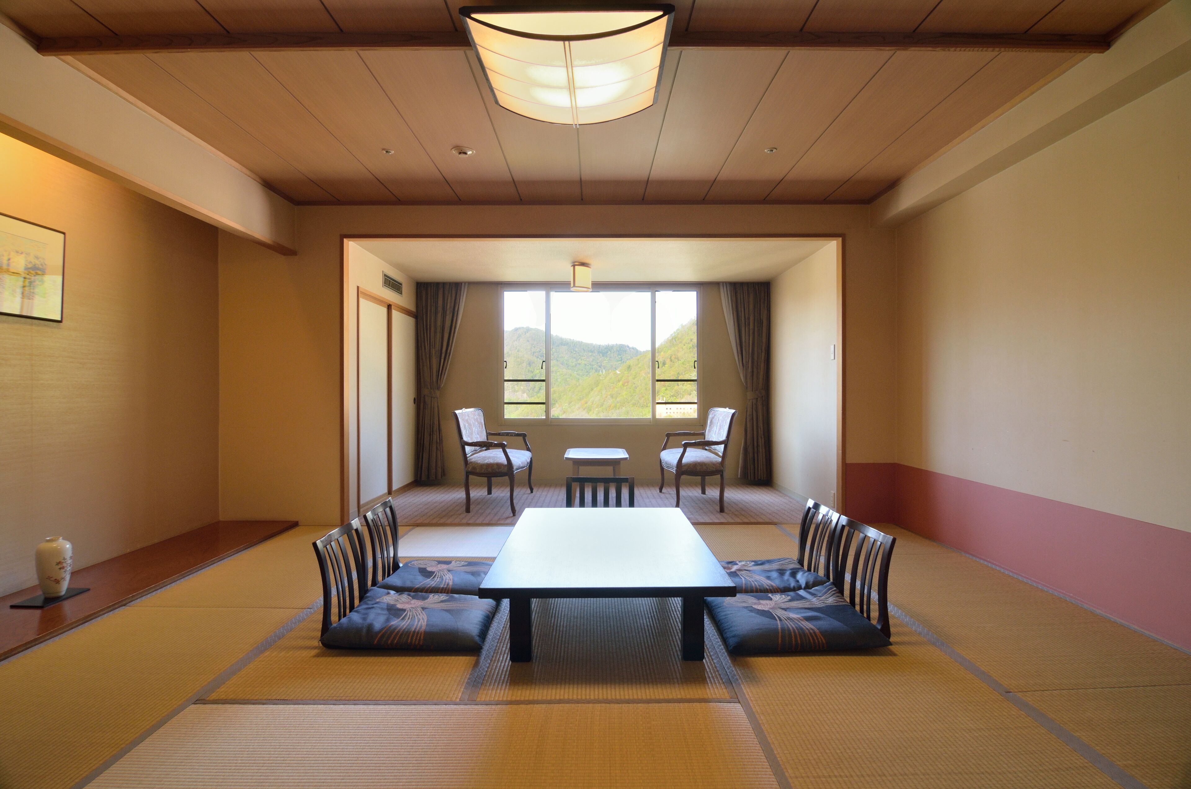 A traditional tatami room with a low table and benches, a large window with a view of greenery, and a ceiling light fixture.