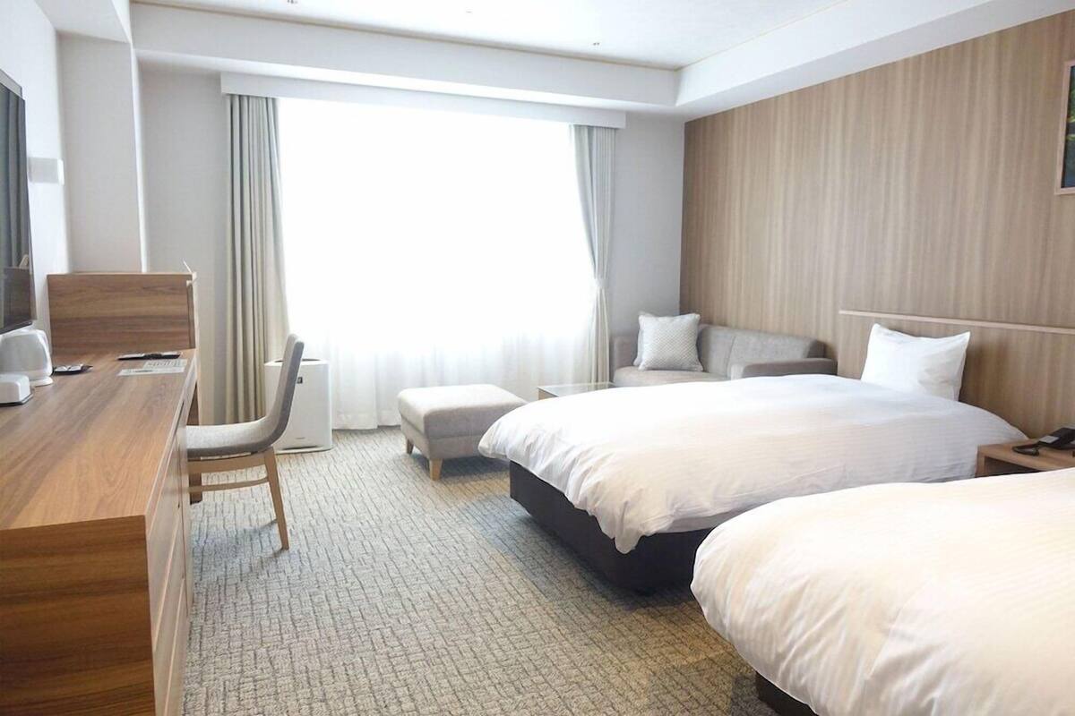 Ein Hotelzimmer mit Schreibtisch, zwei Betten, einem Sessel, einem Fernseher und einem großen Fenster mit Vorhängen.